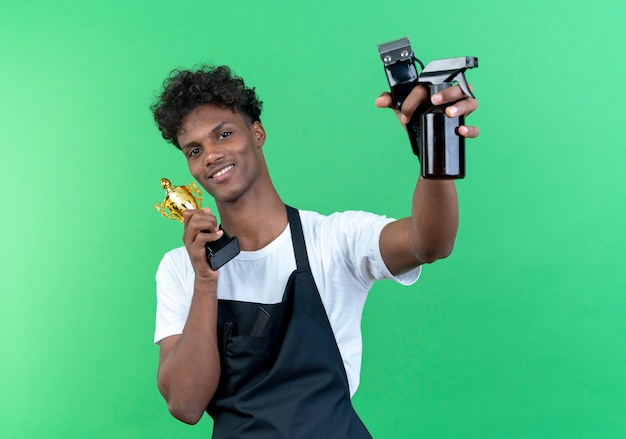 Uśmiechnięty młody afro-amerykański męski fryzjer ubrany w mundur trzymając kubek zwycięzcy wokół twarzy i podnosząc narzędzia fryzjerskie na białym tle na zielonej ścianie