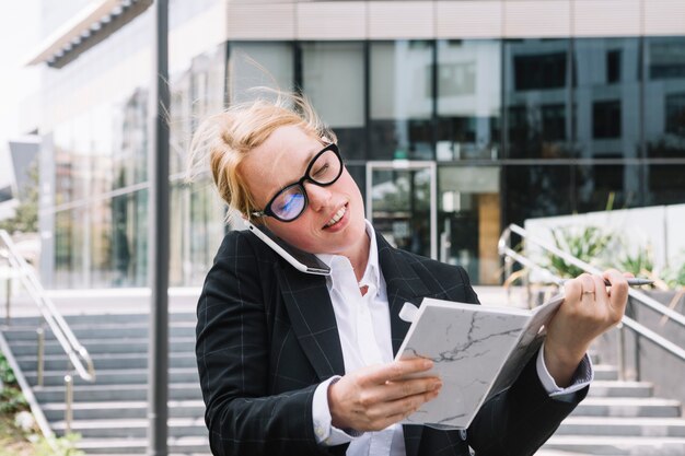 Uśmiechnięty młody bizneswoman opowiada na telefonu komórkowego mienia dzienniczku w ręce