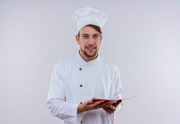 Uśmiechnięty młody brodaty mężczyzna kucharz ubrany w biały mundur kuchenki i kapelusz gospodarstwa notebooka, patrząc na białej ścianie