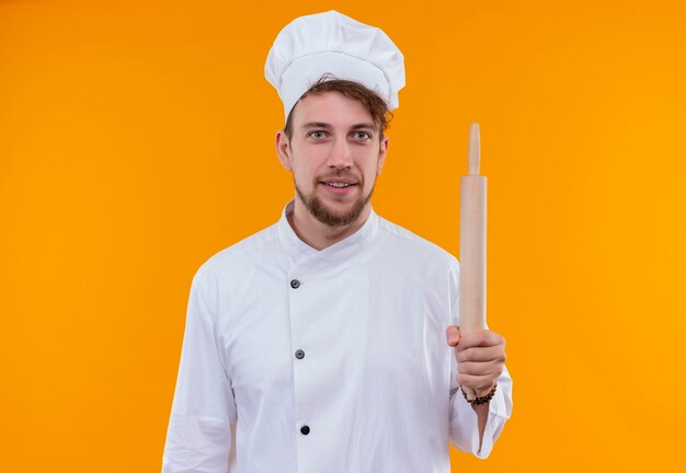 Uśmiechnięty młody brodaty mężczyzna kucharz w białym mundurze trzymając wałek do ciasta, patrząc na pomarańczową ścianę