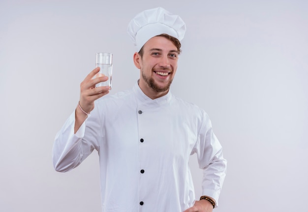 Uśmiechnięty młody brodaty szef kuchni ubrany w biały mundur kuchenki i kapelusz pokazujący szklankę wody, patrząc na białej ścianie