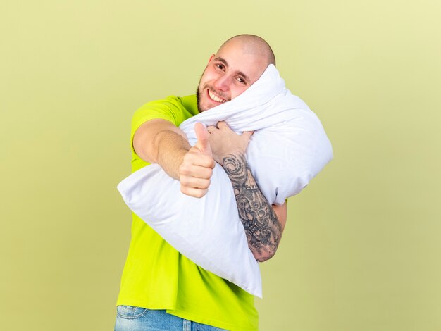 Uśmiechnięty młody chory mężczyzna trzyma poduszkę i kciuki do góry na białym tle na oliwkowej ścianie