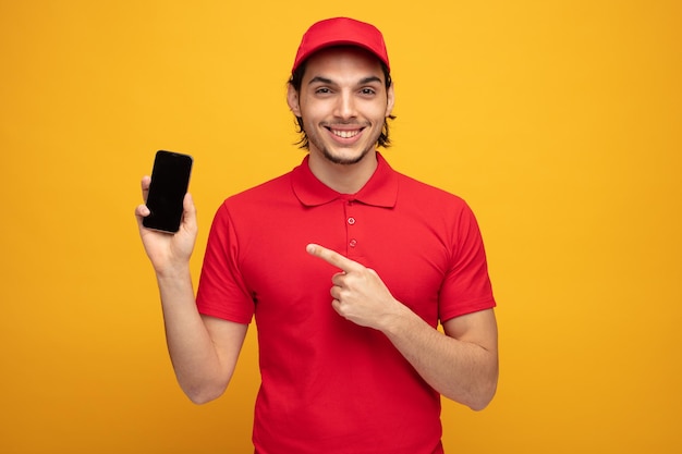 uśmiechnięty młody człowiek dostawy ubrany w mundur i czapkę, patrzący na kamerę pokazującą telefon komórkowy wskazujący na niego na białym tle na żółtym tle