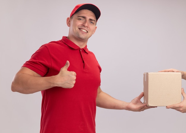Uśmiechnięty młody człowiek dostawy ubrany w mundur z czapką, dając pudełko do klienta pokazując kciuk do góry na białym tle na białej ścianie
