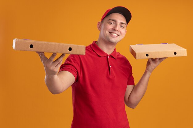 Uśmiechnięty młody człowiek dostawy ubrany w mundur z czapką, trzymając pudełka po pizzy na białym tle na pomarańczowej ścianie