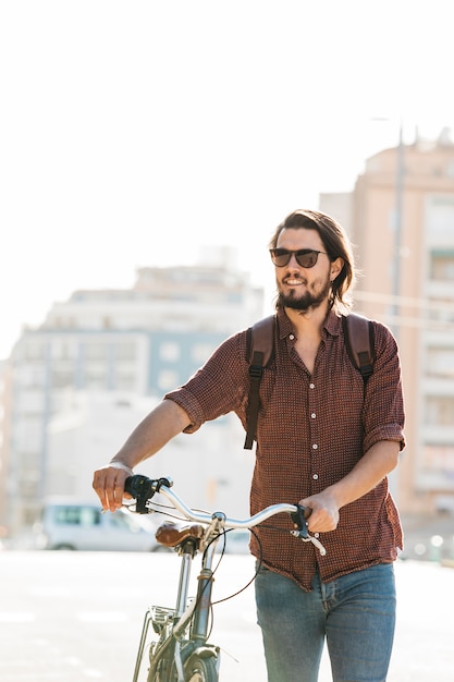 Uśmiechnięty młody człowiek jest ubranym okulary przeciwsłonecznych chodzi z bicyklem na drodze w popołudniu