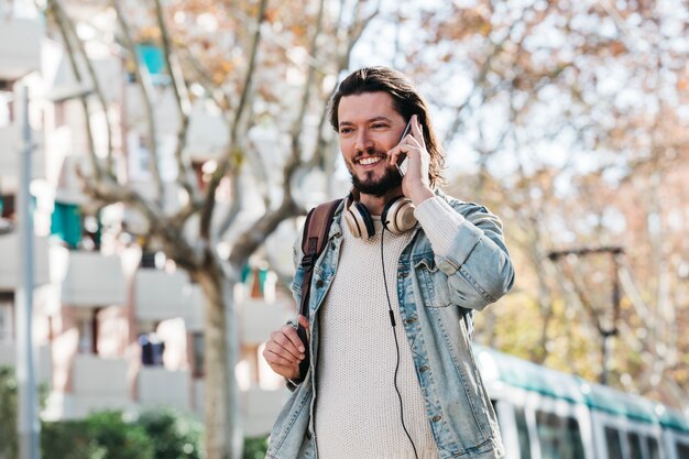 Uśmiechnięty młody człowiek opowiada na telefonie komórkowym przy outdoors z jego plecakiem