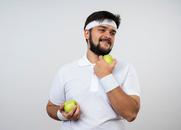 Uśmiechnięty młody człowiek sportowy patrząc na bok noszenie opaski i nadgarstka, trzymając jabłka na białym tle na białej ścianie