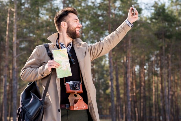 Uśmiechnięty młody człowiek trzyma mapę w ręku biorąc selfie w lesie z telefonu komórkowego