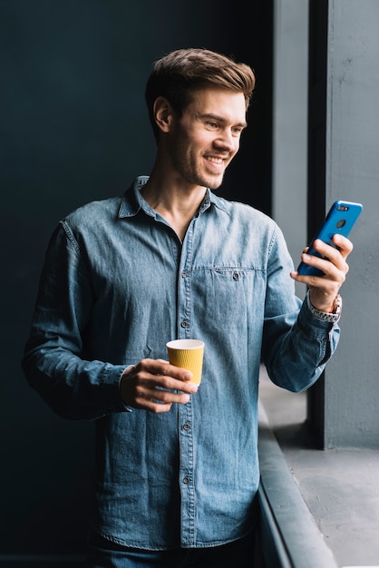 Uśmiechnięty Młody Człowiek Trzyma Takeaway Filiżankę Patrzeje Telefon Komórkowego