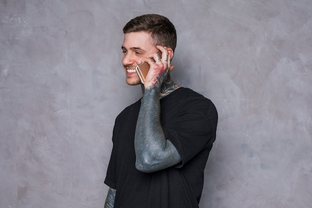 Uśmiechnięty młody człowiek z tatuażem na ręce rozmawia przez telefon komórkowy na szarej ścianie