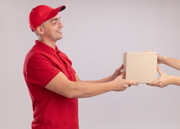 Uśmiechnięty młody dostawca w mundurze z czapką dającą pudełko klientowi na białej ścianie