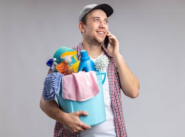 uśmiechnięty młody facet sprzątający w czapce trzymającej wiadro z narzędziami do czyszczenia mówi przez telefon odizolowany na białej ścianie