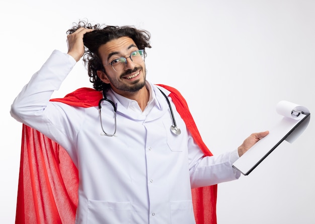 Uśmiechnięty młody kaukaski mężczyzna w okularach optycznych w mundurze lekarza z czerwonym płaszczem i stetoskopem na szyi podnosi włosy ręką i trzyma schowek na białej ścianie