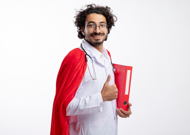 Uśmiechnięty młody kaukaski mężczyzna w okularach optycznych w mundurze lekarza z czerwonym płaszczem i stetoskopem na szyi stoi bokiem, trzymając folder z plikami i kciuki w górę z miejscem na kopię