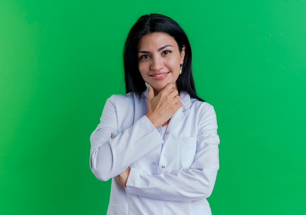 Uśmiechnięty młody lekarz kobiet na sobie szatę medyczną dotykając podbródka na białym tle na zielonej ścianie z miejsca na kopię