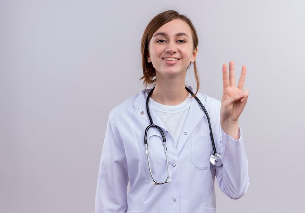 Uśmiechnięty młody lekarz kobiet na sobie szatę medyczną i stetoskop pokazujący trzy na izolowanych białej ścianie z miejsca na kopię