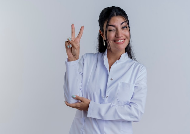 Uśmiechnięty młody lekarz kobiet na sobie szatę medyczną robi znak pokoju kładąc rękę na łokciu na białym tle na białej ścianie z miejsca na kopię