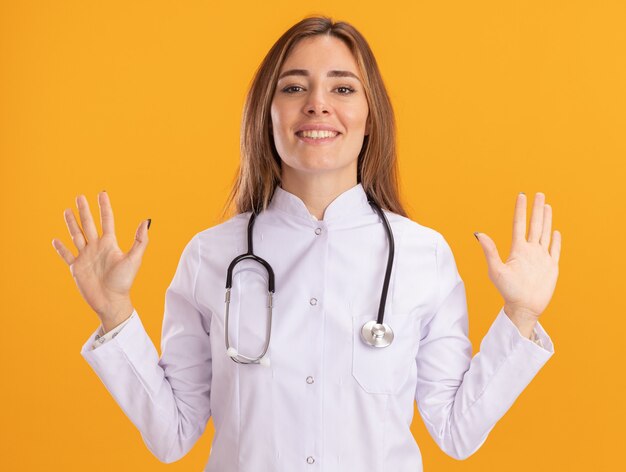 Uśmiechnięty młody lekarz kobiet na sobie szatę medyczną ze stetoskopem, rozkładając ręce na białym tle na żółtej ścianie
