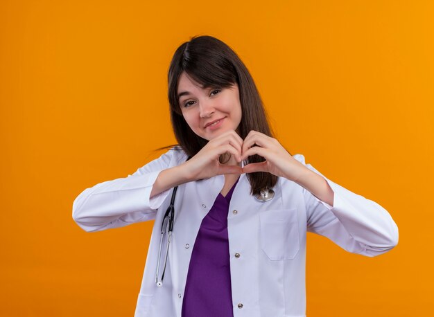 Uśmiechnięty młody lekarz kobiet w szatę medyczną z serca gesty stetoskop obiema rękami na na białym tle pomarańczowym tle z miejsca na kopię