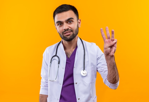 Uśmiechnięty młody lekarz mężczyzna ma na sobie stetoskop medycznej sukni trzy na odosobnionym żółtym tle