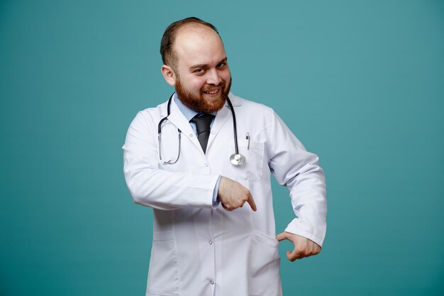 Uśmiechnięty młody lekarz mężczyzna ubrany w fartuch medyczny i stetoskop na szyi, patrzący na kamerę wskazującą na jego kieszeń na białym tle na niebieskim tle