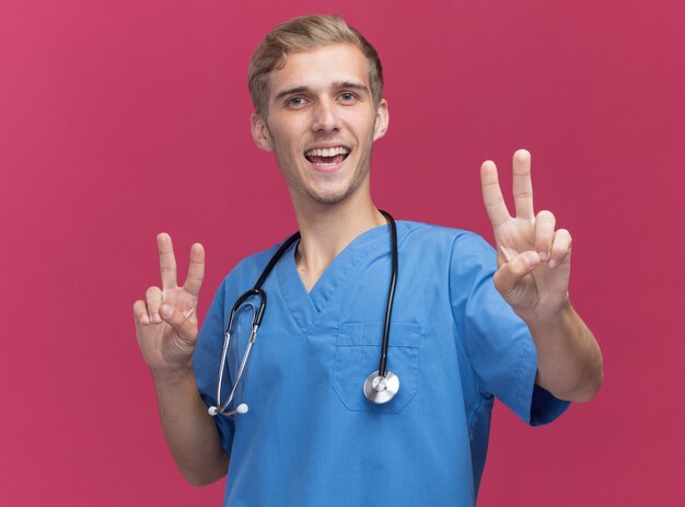Uśmiechnięty młody lekarz mężczyzna ubrany w mundur lekarza ze stetoskopem pokazujący gest pokoju na białym tle na różowej ścianie