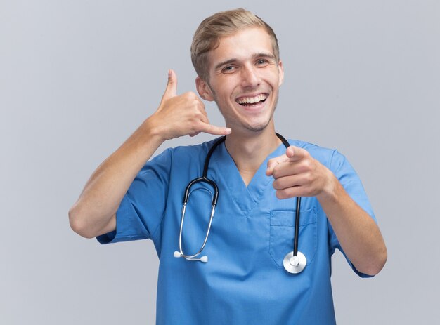 Uśmiechnięty młody lekarz mężczyzna ubrany w mundur lekarza ze stetoskopem pokazujący gest połączenia telefonicznego i punkty na białym tle na białej ścianie