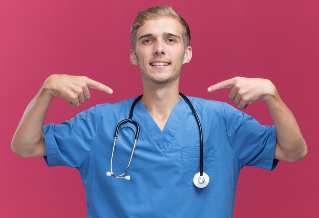 Uśmiechnięty młody lekarz mężczyzna ubrany w mundur lekarza ze stetoskopem wskazuje na siebie na białym tle na różowej ścianie