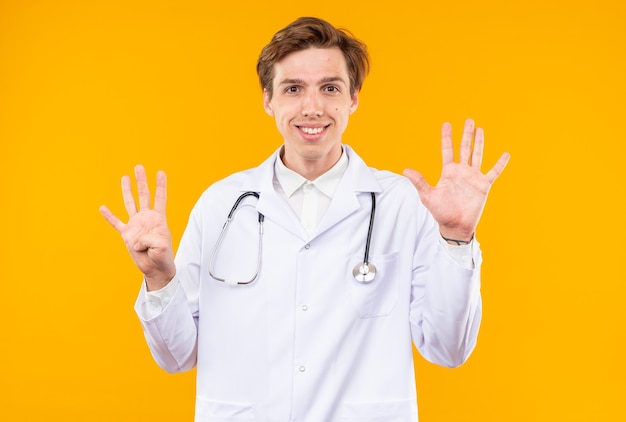 uśmiechnięty młody lekarz mężczyzna ubrany w szatę medyczną ze stetoskopem pokazujący różne liczby na pomarańczowej ścianie