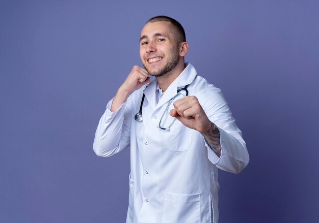 Uśmiechnięty młody lekarz mężczyzna ubrany w szlafrok medyczny i stetoskop robi gest boksu z przodu na białym tle na fioletowej ścianie