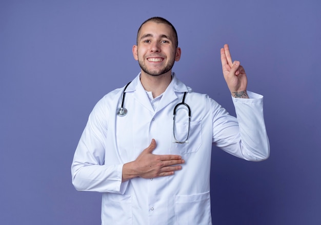 Uśmiechnięty młody lekarz mężczyzna ubrany w szlafrok medyczny i stetoskop robi gest obietnicy na białym tle na fioletowej ścianie