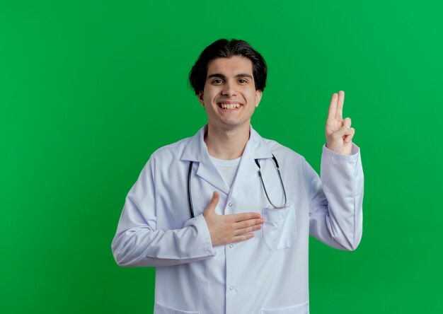 Uśmiechnięty młody lekarz mężczyzna ubrany w szlafrok medyczny i stetoskop robi gest obietnicy na białym tle na zielonej ścianie z miejsca na kopię