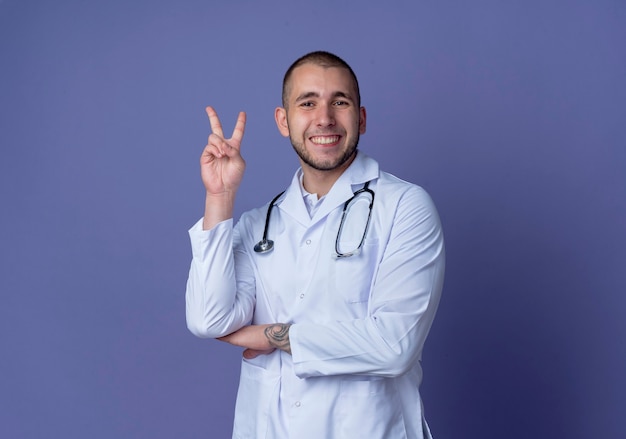 Uśmiechnięty młody lekarz mężczyzna ubrany w szlafrok medyczny i stetoskop robi znak pokoju kładąc rękę pod łokciem na białym tle na fioletowej ścianie