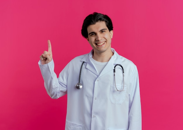 Bezpłatne zdjęcie uśmiechnięty młody lekarz mężczyzna ubrany w szlafrok medyczny i stetoskop skierowany w górę na białym tle na różowej ścianie z miejsca na kopię