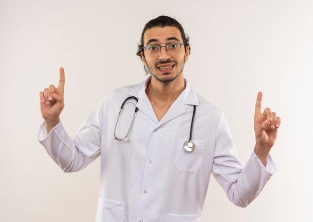 Uśmiechnięty młody lekarz mężczyzna z okularami optycznymi na sobie białą szatę z punktami stetoskopu do góry na odosobnionej białej ścianie z miejsca na kopię
