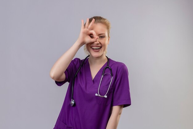Uśmiechnięty młody lekarz ubrany w fioletową medyczną suknię i stetoskop pokazuje wygląd gestu na odizolowanej białej ścianie