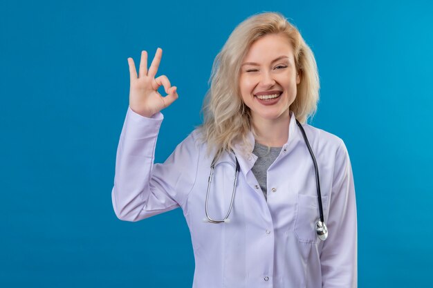 Uśmiechnięty młody lekarz ubrany w stetoskop w sukni medycznej pokazujący gest okey na niebieskiej ścianie