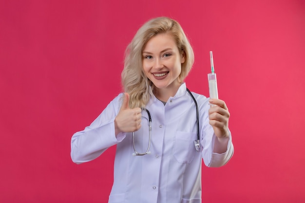 Uśmiechnięty młody lekarz ubrany w stetoskop w sukni medycznej trzymając strzykawkę jej kciuk na czerwonym backgroung