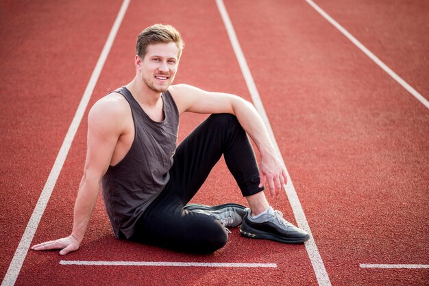 Uśmiechnięty młody męski atleta siedzi na czerwonym biegowym śladzie blisko linii początkowej
