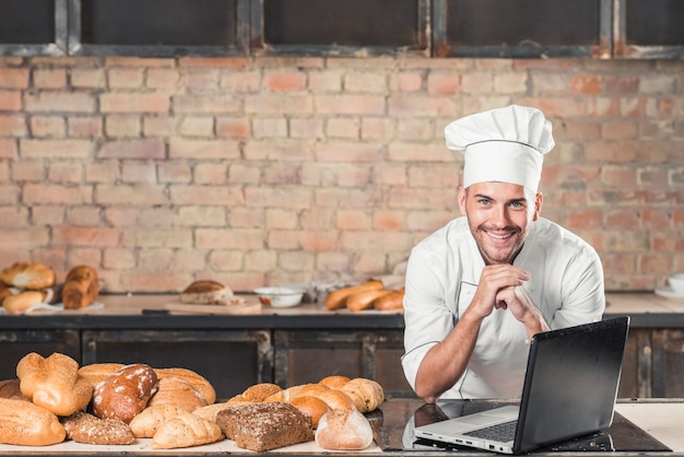 Uśmiechnięty młody męski piekarz opiera na stole z rozmaitością piec chleb i laptop