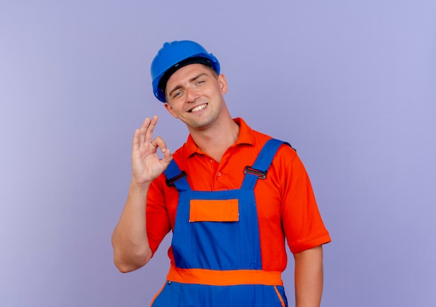 Uśmiechnięty młody mężczyzna budowniczy na sobie mundur i kask pokazujący okey gest na fioletowo