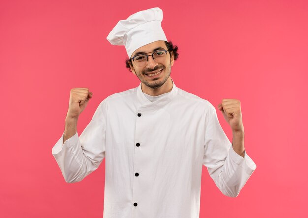 Uśmiechnięty młody mężczyzna kucharz na sobie mundur szefa kuchni i okulary pokazujące tak gest na białym tle na różowej ścianie