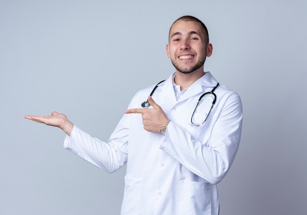 Uśmiechnięty młody mężczyzna lekarz ubrany w medyczny szlafrok i stetoskop na szyi, pokazując pustą rękę i wskazując na nią na białym tle na białej ścianie