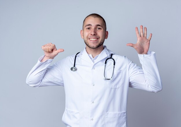 Uśmiechnięty młody mężczyzna lekarz ubrany w medyczny szlafrok i stetoskop na szyi, pokazując sześć z rękami odizolowanymi na białej ścianie