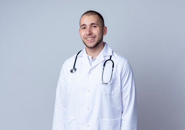 Uśmiechnięty młody mężczyzna lekarz ubrany w medyczny szlafrok i stetoskop na szyi, stojąc i patrząc z przodu na białym tle na białej ścianie