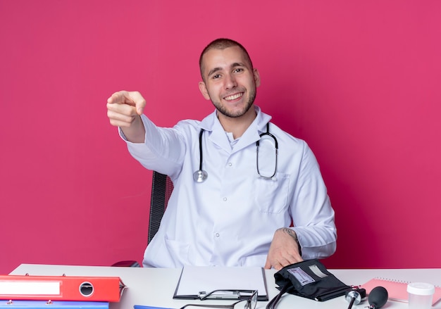 Uśmiechnięty Młody Mężczyzna Lekarz Ubrany W Szlafrok Medyczny I Stetoskop Siedzi Przy Biurku Z Narzędziami Pracy, Wskazując Prosto Na Białym Tle Na Różowej ścianie