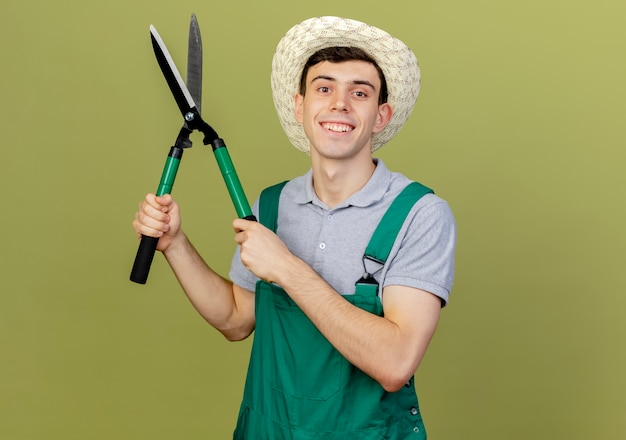 Uśmiechnięty młody mężczyzna ogrodnik w kapeluszu ogrodniczym patrzy na aparat trzymający maszynki do strzyżenia