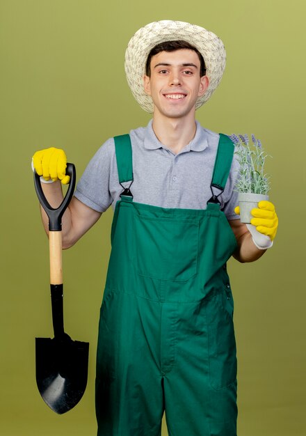 Uśmiechnięty młody mężczyzna ogrodnik w kapeluszu ogrodniczym trzyma szpadel i kwiaty w doniczce