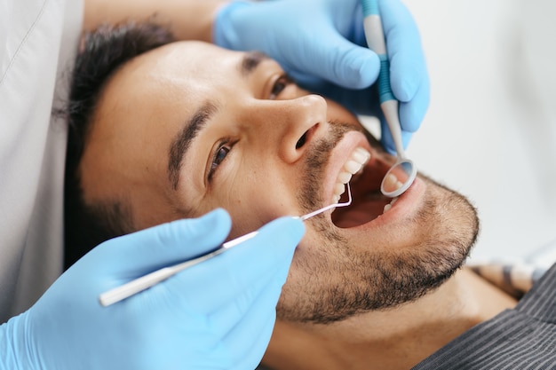 Uśmiechnięty młody mężczyzna siedzi na fotelu dentysty, podczas gdy lekarz bada jego zęby